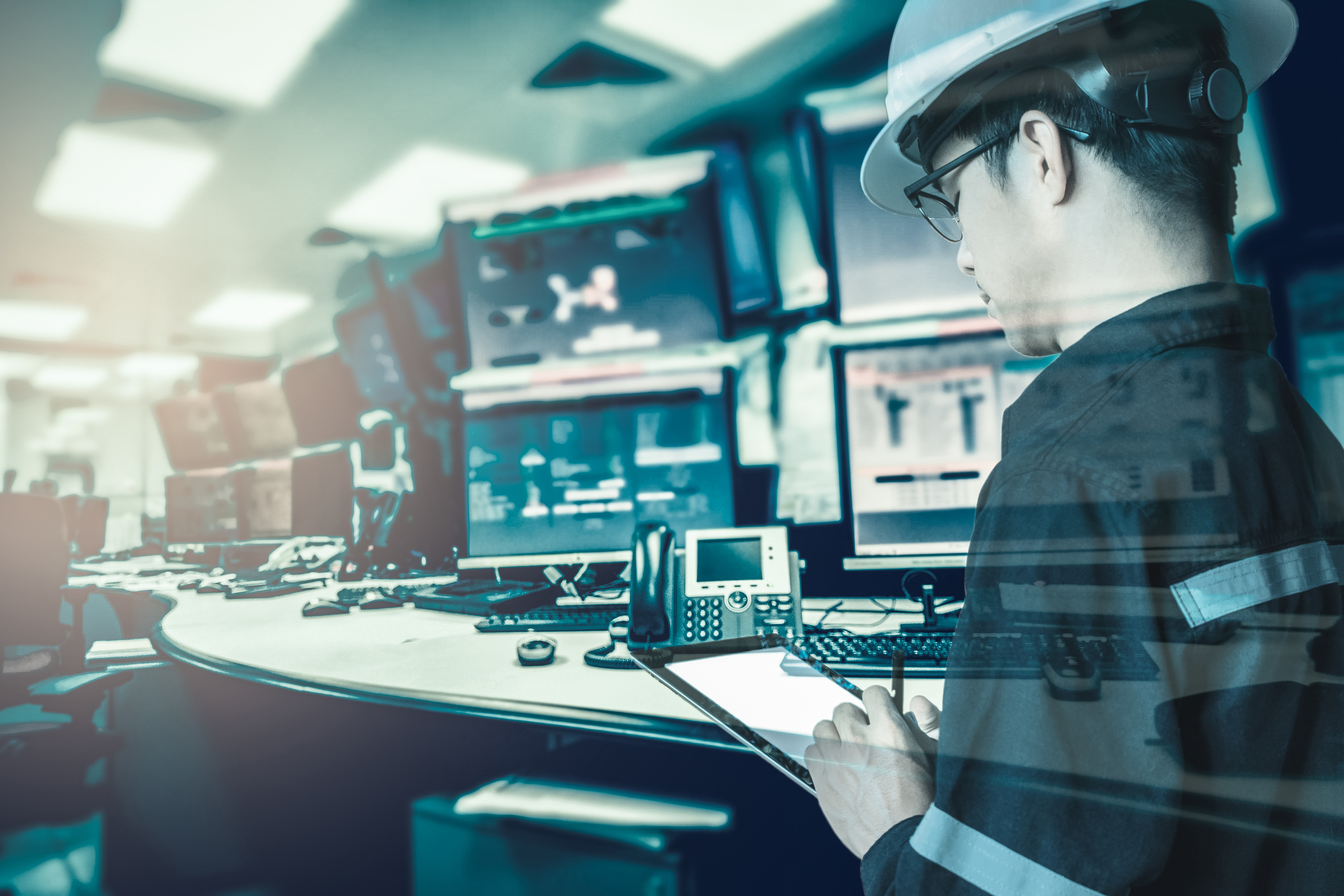 A man in a hardhat is holding a tablet and looking and several computer screens.